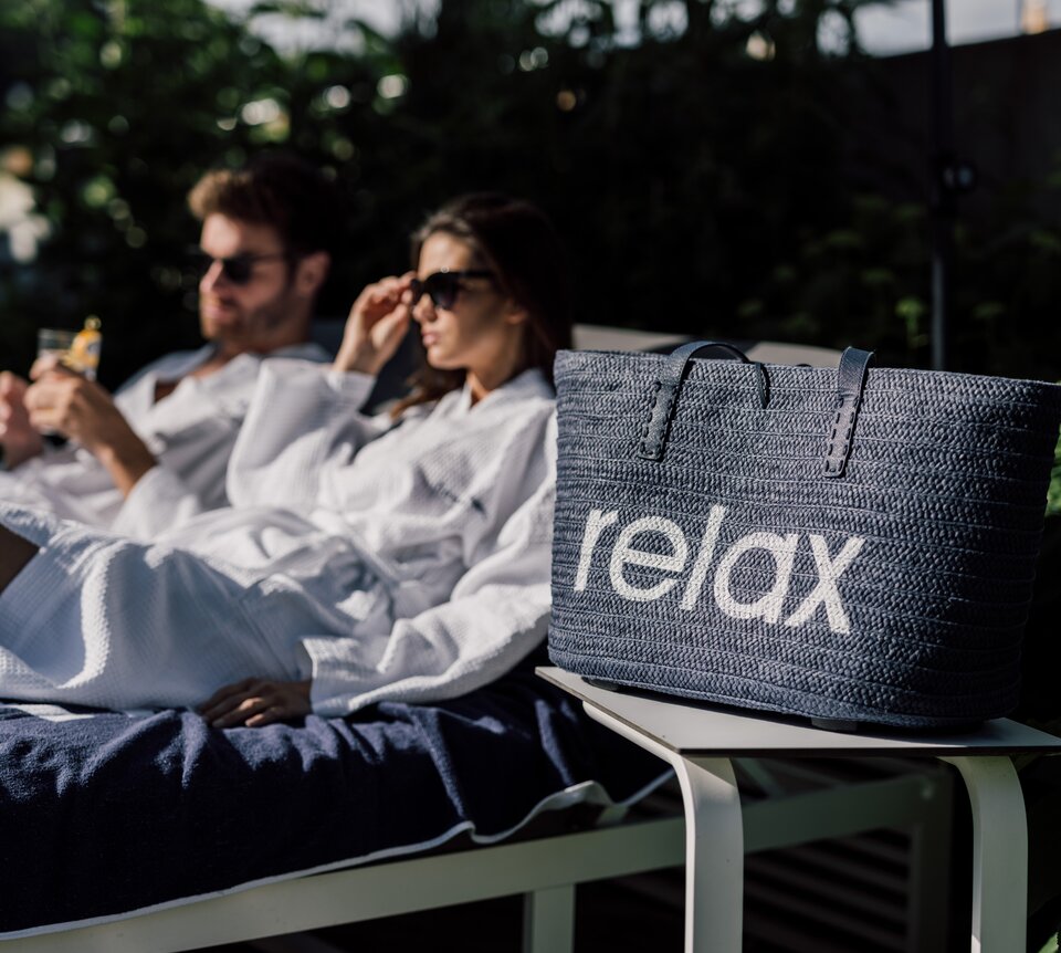 Paar entspannt auf einer Liege im Relax! Garten, daneben eine Relax! Badetasche | © Spa Resort Geinberg / Chris Perkles