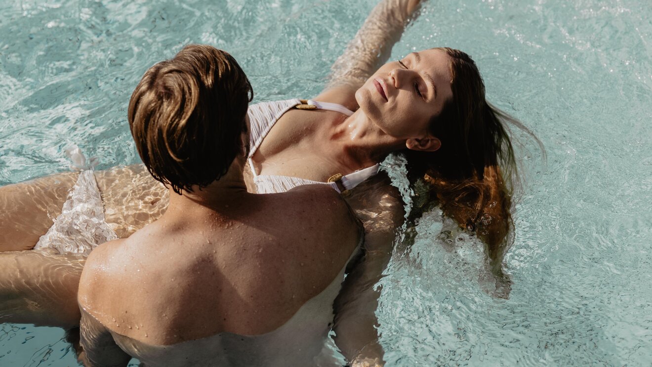 Mann trägt blonde Frau auf Händen im Wasser des Thermal-Beckens outdoor im Spa Resort Geinberg | © Spa Resort Geinberg / Chris Perkles