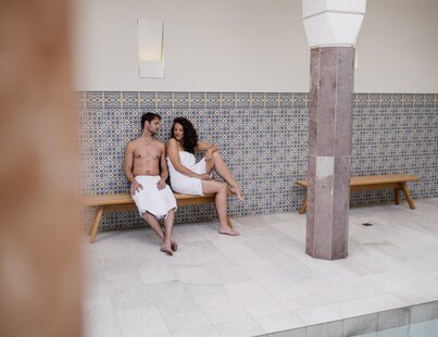 Paar im Pestemaltuch sitzt auf einer Bank im Arkadenhof der Oriental World im Spa Resort Geinberg | © Spa Resort Geinberg / Chris Perkles